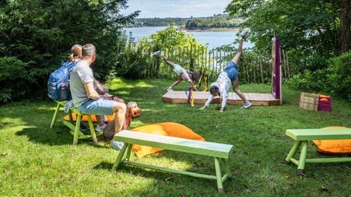 A family sitting on colourful benches surrounding an outdoor stage area where their children dance during the summer with views across the Menai Strait behind